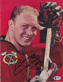 The Bobby Hull Signed Chicago Blackhawks Magazine Page Photo by Sports Integrity features Hull in a red jersey, holding a hockey stick with gloves. The green, yellow, and white logo is visible, and his black signature appears across his chest on the red background.