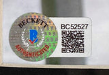 Close-up of a Beckett authentication sticker with holographic seal, AUTHENTICATED text, blue “B” logo, serial BC52527, and QR code on white—on Johnny Depp Signed Framed 8x10 Pirates Of The Caribbean Photo by Sports Integrity.
