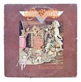 The image features the Aerosmith Toys in the Attic 1975 Vinyl Record 1 by Sports Integrity, showing the band’s logo and an illustration of old toys, including dolls and a horse, coming to life in a dusty attic.