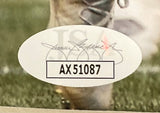 A close-up of a JSA-authenticated Amon-Ra St. Brown signed Detroit Lions vs Seattle 8x10 photo by Sports Integrity, showing an oval sticker with signature and code AX51087, placed on a sports surface with a cleat in the background.