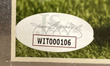A white oval JSA hologram on a Calvin Johnson Signed Framed 8x10 Detroit Lions Photo by Sports Integrity, with the code WIT000106 in red-bordered box and faint signatures, placed on a surface with green grass behind.