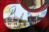 A close-up of a Dan & Shay Signed 41" Bigger Houses Red Acoustic Guitar by Sports Integrity, featuring a photo of the duo on a roof with guitars, signed in black marker and a visible QR code label beneath the image.