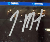 A close-up of a Ja Morant Signed Framed 8x10 Grizzlies vs Timberwolves Photo by Sports Integrity shows his silver autograph on the glass, with blurred lights and blue FanDuel signs in the background.