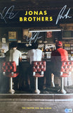 Three people sit on red barstools at a vintage diner counter as seen from behind, with "Jonas Brothers" and their signatures above. Below is the text: "The Poster For The Album." Product: Jonas Brothers Signed 11x17 The Album Poster by Sports Integrity.
