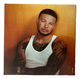Kane Brown poses with arms crossed, showing tattoos and a white tank top, for Sports Integrity’s signed "The High Road" CD booklet (PSA/DNA), against an orange-brown background.
