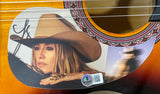 A close-up of the Sports Integrity Lainey Wilson Signed 38" Acoustic Guitar BAS shows a decal with a woman in a cowboy hat, chin on hand, a blurred cowboy in the background, plus her signature and a QR code.