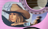 A close-up of a woman's face in a cowboy hat appears on the Lainey Wilson Signed 38" Pink Acoustic Guitar by Sports Integrity, featuring western designs and a visible autograph. A blurred figure in a hat is also nearby.