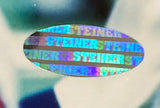 Oval-shaped holographic Sports Integrity sticker with "STEINER" repeatedly printed in blue and green, included on the Phil Simms Signed Framed 8x10 New York Giants Photo (Steiner Sports Hologram).