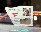 A close-up of a Sports Integrity sticker with code “KF50462200,” a QR code, red logo, and blue “M” on the Rhea Ripley Signed Framed 8x10 WWE Wrestlemania 39 Photo. The background is blurry with reflections and shapes.