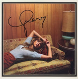 A woman with light brown hair relaxes on a retro couch in a wood-paneled room, her arm on her forehead. Beside her, a table holds the Sports Integrity Taylor Swift Signed Framed Insert w/ Midnights Vinyl BAS AE02656.