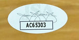 A white oval sticker with black code AC65303 and a faded signature is affixed to the Tyler Herro Signed Framed 16x20 Miami Heat Photo JSA from Sports Integrity, shown against a brown background.