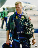 The Sports Integrity Val Kilmer Signed 16x20 Top Gun Photo (JSA) shows a young male fighter pilot in a green flight suit holding a helmet outdoors, with aircraft and crew members blurred in the background on an airstrip.